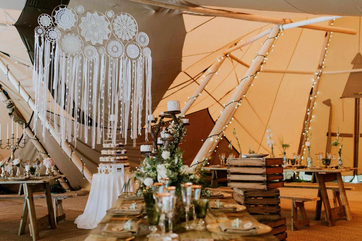 A rustic and stylish wedding setting, in a teepee, with wooden beams, a large dream catcher, glass and wooden place settings on a long oak bench. Apple crates, fairy lights and greenery in the background.